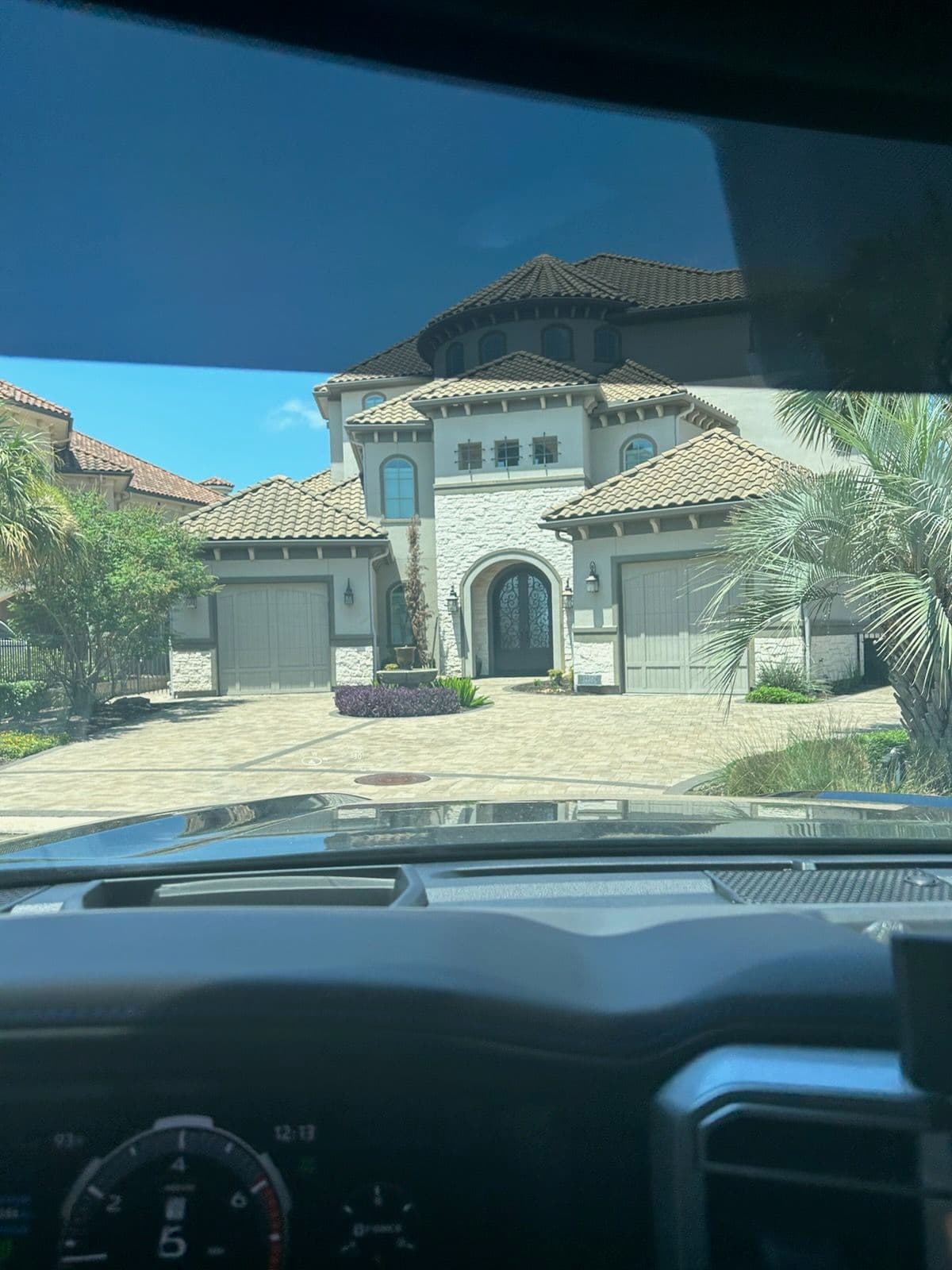 View of a luxury home with a tiled roof and palm trees from inside a vehicle.